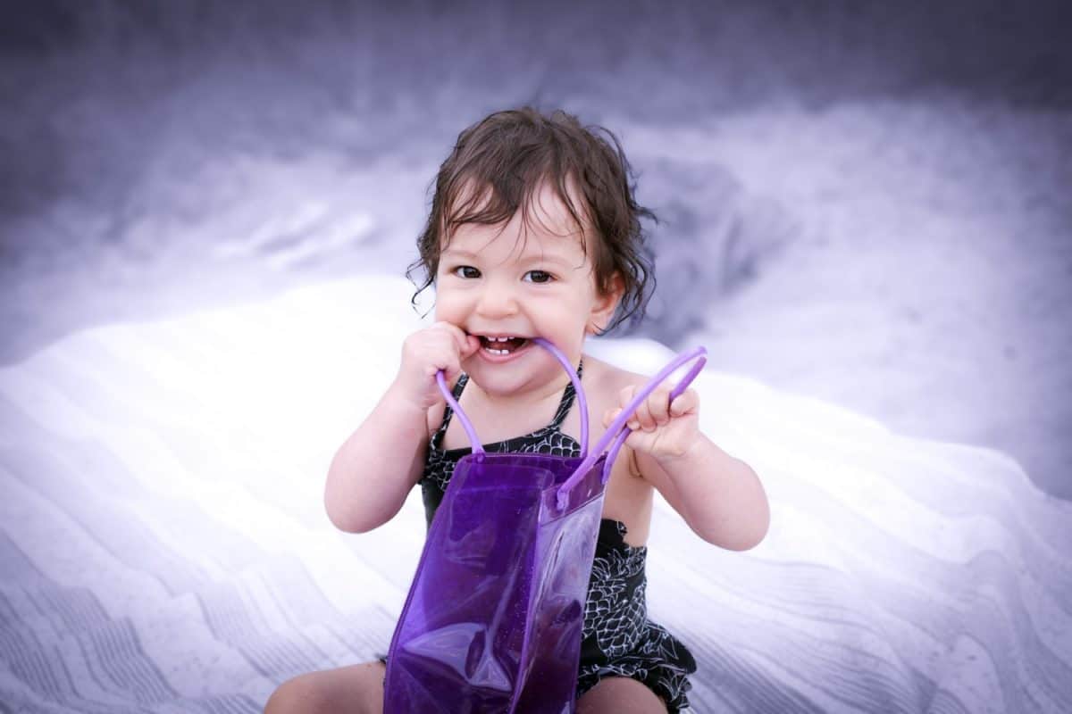 Cute teething baby girl with brown hair and eyes in the swimsuit smiling and chewing on a purple plastic bag handle while sitting outside on a blanket in the beach on a blurry lilac background