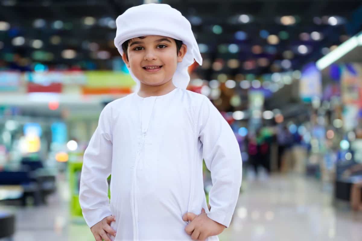 Young Arab boy inside a shopping mall. Emirati kid wearing Kandura traditional clothing for Middle East concept with retail shop as background. Arabic son dishdasha.