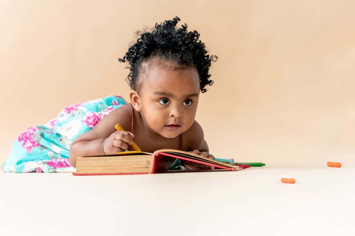 A 2-year-old Nigerian baby girl with beautiful curly hair, sitting on the floor, holding a yellow crayon and looking up. African baby and enhancing the development of children by studying art concept.