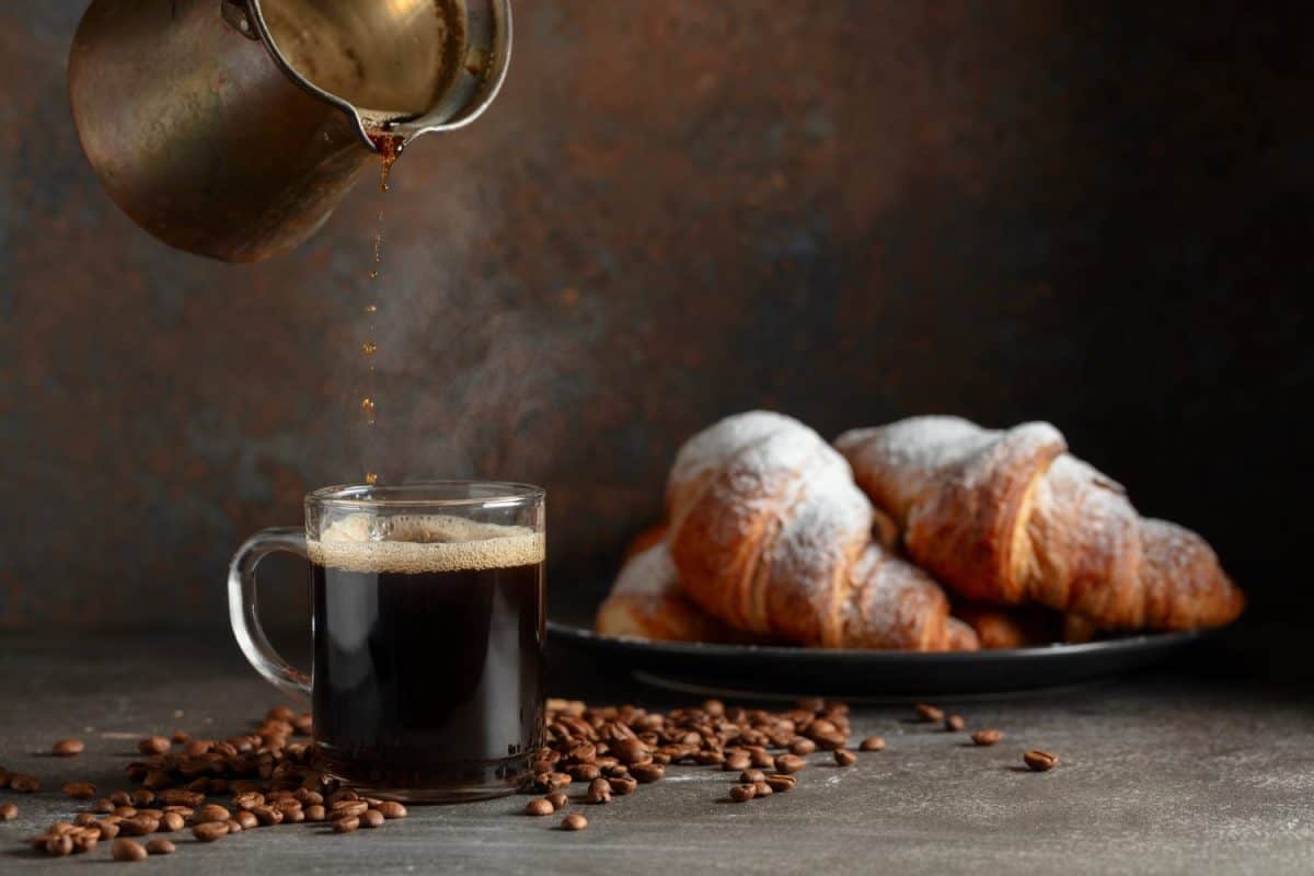 Coffee is poured into a mug. Croissants and black coffee on a kitchen table. Focus on a foreground.