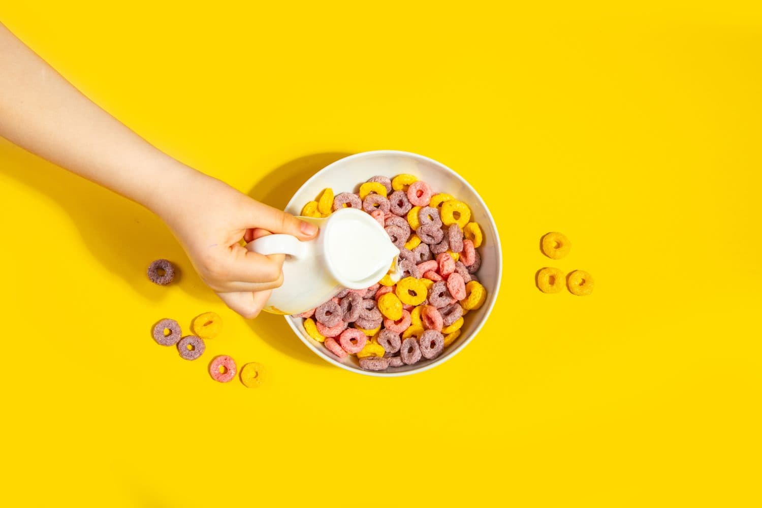 Bowl of colorful cereal corn rings on yellow table. Breakfast concept. Top view. Child kid hand poring milk.