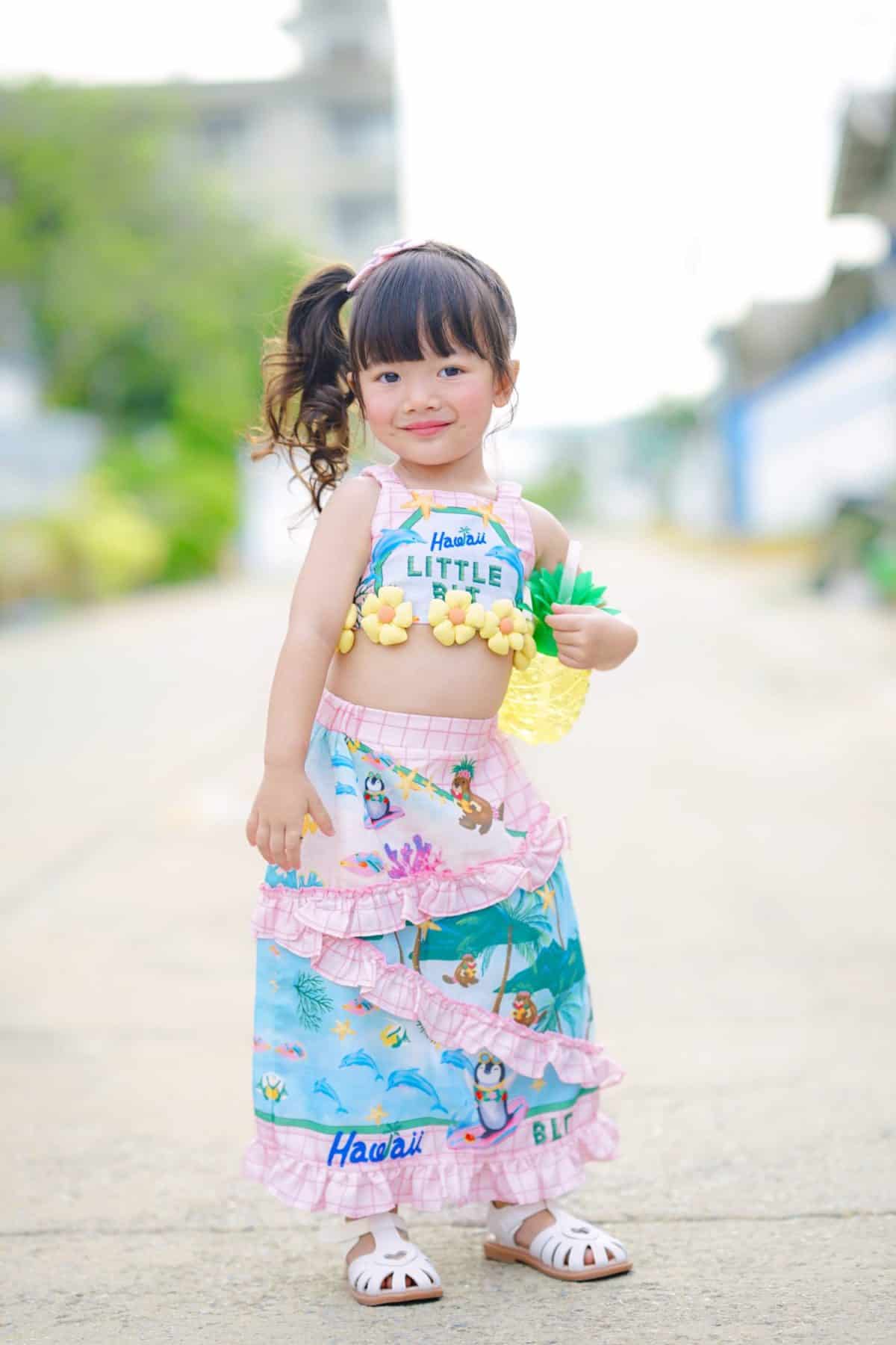 The long-haired girl in a crop top and high-waisted skirt with flower patterns, styled in a Hawaiian fashion, is posing for a photo with a cheerful, lively, and playful demeanor.