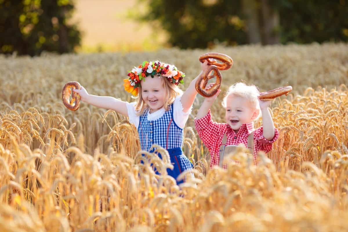 Child in wheat field with bread and pretzel. Oktoberfest time in Germany. Kids in traditional German dirndl dress and leather pants in autumn harvest crop field.