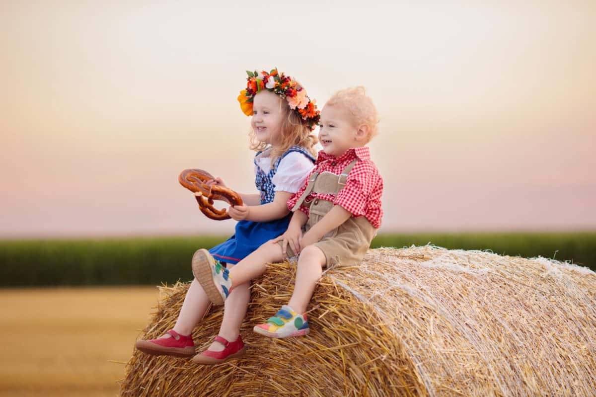 Child in wheat field with bread and pretzel. Oktoberfest time in Germany. Kids in traditional German dirndl dress and leather pants in autumn harvest crop field.