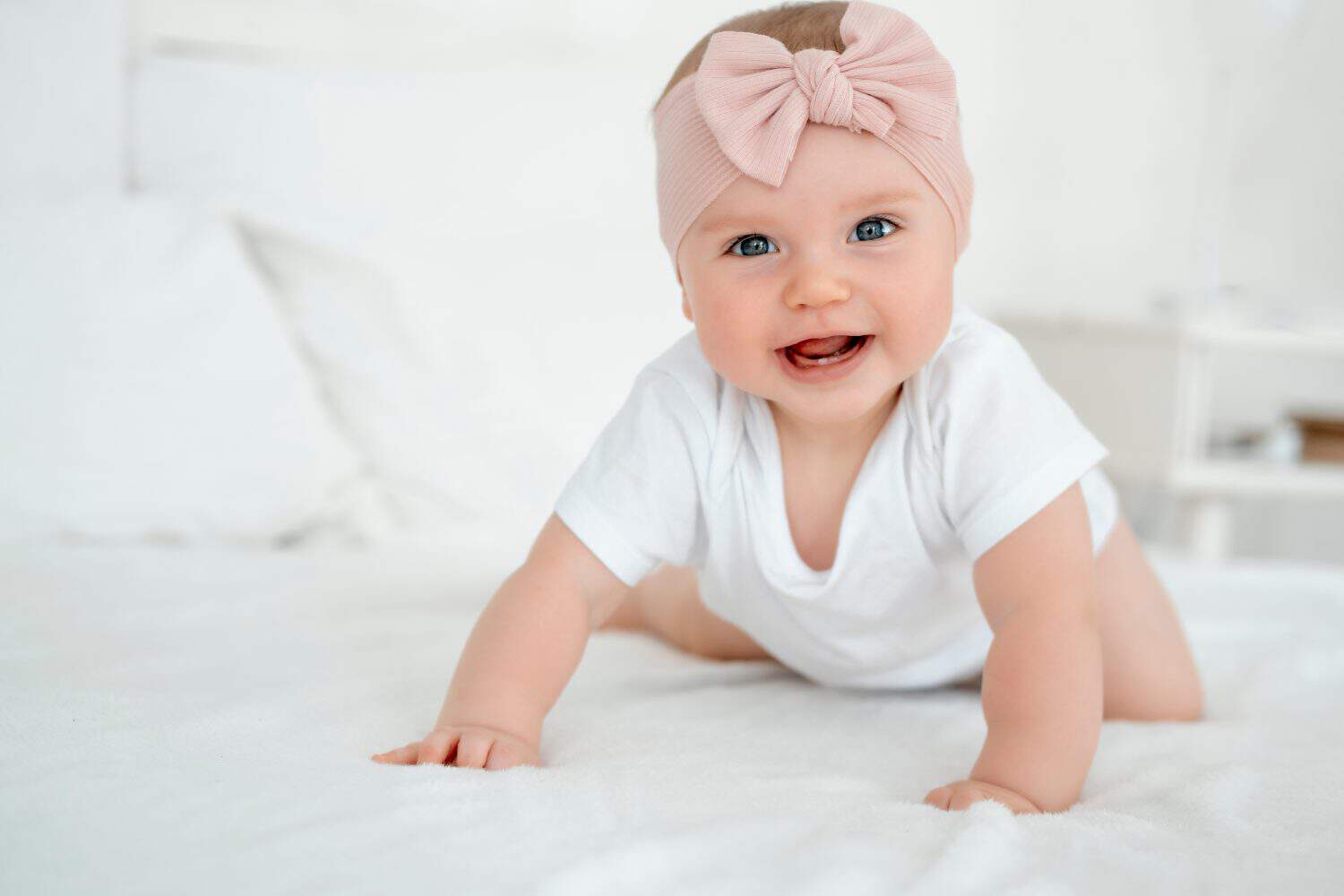 A baby girl is smiling at home on a white bed with a bow on her head, a happy healthy child of six months is laughing.