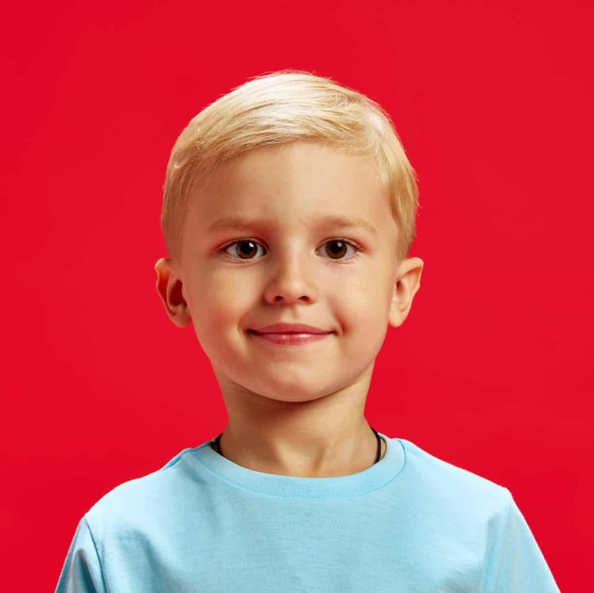 Portrait of adorable little baby boy, blond haired child wearing blue t-shirt and looking at camera with warm smile against red studio background. Concept of childhood, emotions, fashion, lifestyle