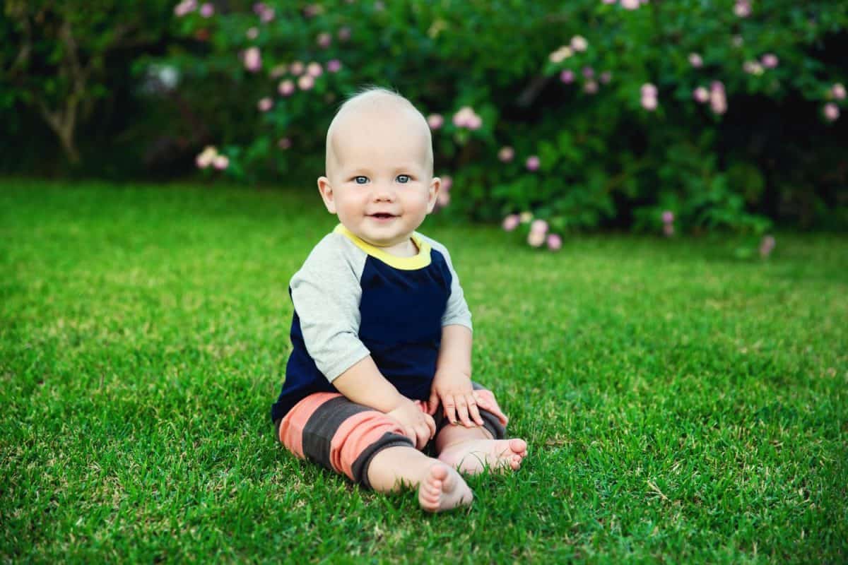 Happy adorable baby boy sitting on the grass and laughing in summer day. Happy smile, 8 months old. Child in trendy and cute clothes