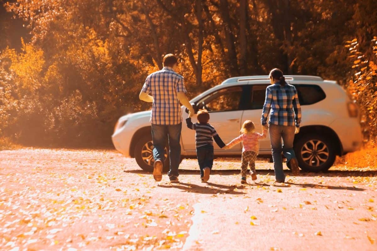 Parents - mother, father and kids - daughter, son holding hands each other against white SUV big car. Outdoor photo in autumn park. Casual dress. Sunset light rays