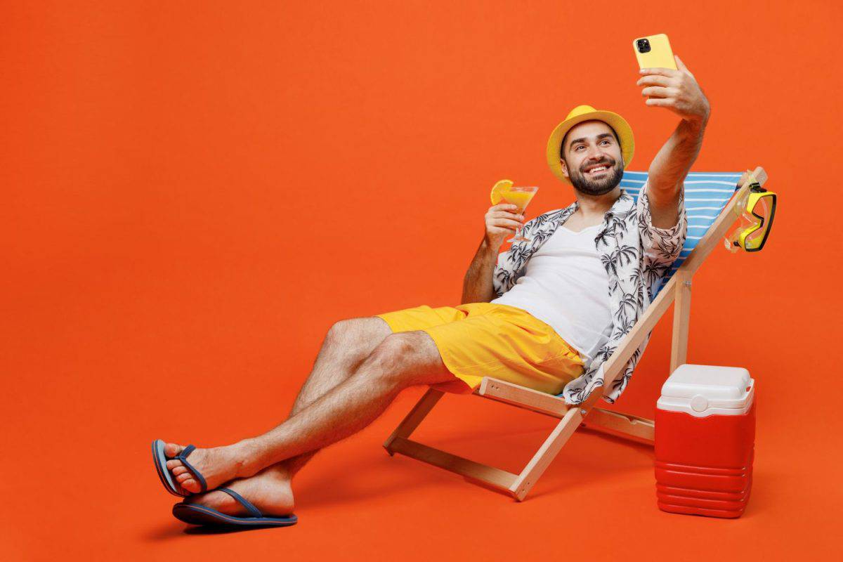 Young tourist man wear beach shirt hat lie on deckchair near fridge drink cocktail do selfie shot on mobile cell phone isolated on plain orange background . Summer vacation sea rest sun tan concept