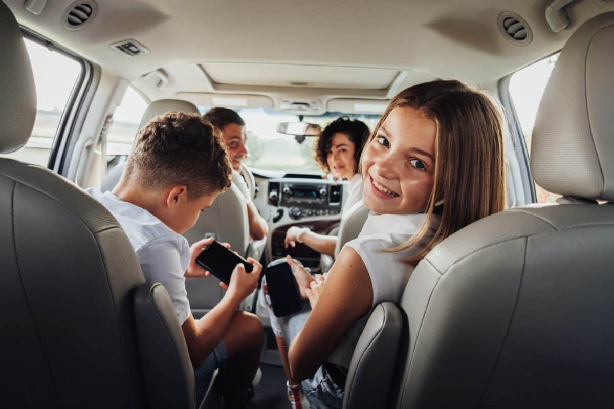 Cheerful Caucasian Teenage Girl Smiles Into Camera While Sitting in Minivan Car with Her Brother, Mother and Father, Happy Four Members Family Enjoying Weekend Road Trip