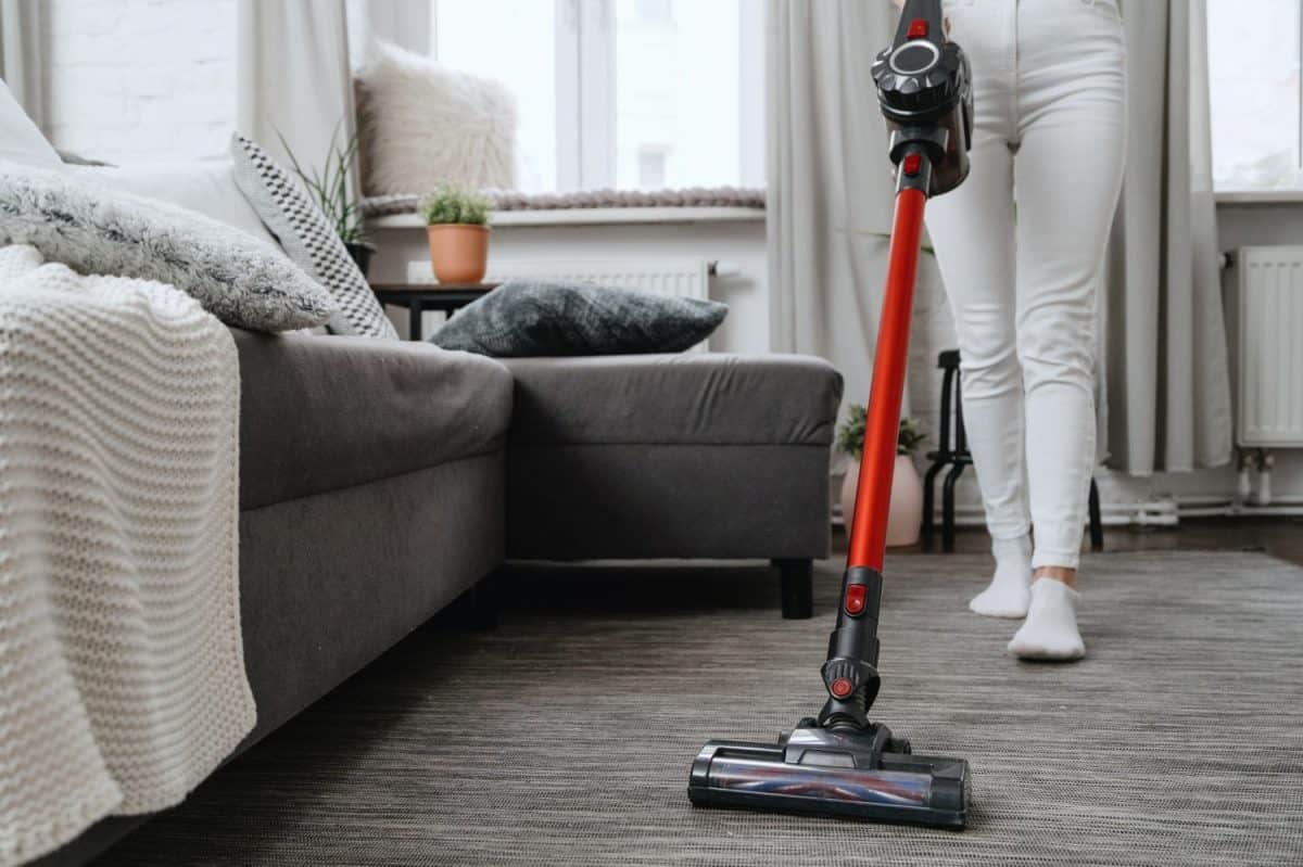 cropped shot of housewife in white jeans using cordless handheld vacuum cleaner in living room with sofa and pillows, household chores concept