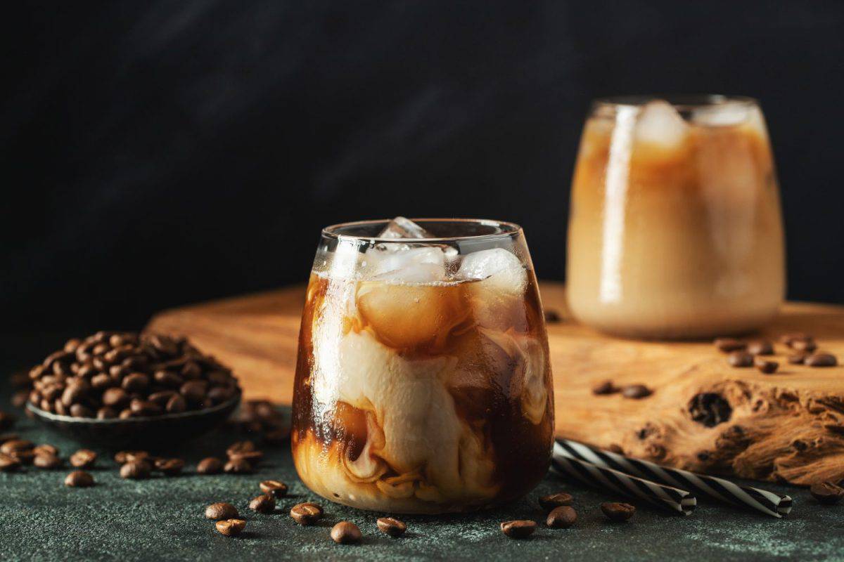 Ice coffee in a tall glass with cream poured over, ice cubes and beans on a dark concrete table.