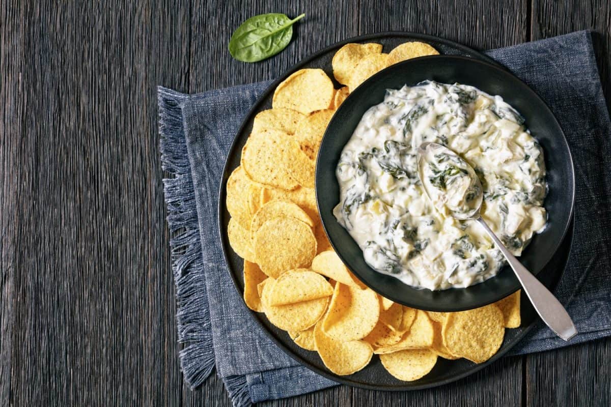 artichoke spinach almond creamy dip with potato chips in bowl on black platter on dark wooden table with ingredients, horizontal view, flat lay, free space. Forget Chick-fil-A Sauce, Americans Go Crazy for These 8 Condiments