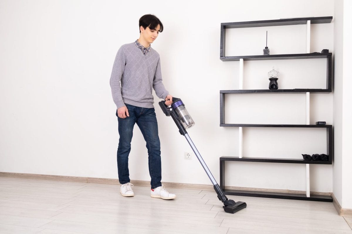 A young man is using a cordless vacuum cleaner to clean a laminate floor in a white room. In the background is a modern bookshelf with decorative objects. I Loved My Dyson But These 8 Vacuum Brands Are a Better Deal