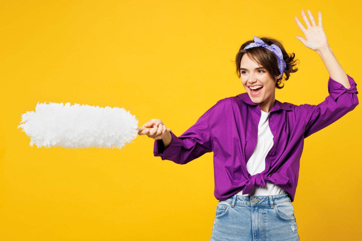 Side profile view young fun woman wear purple shirt do housework tidy up hold duster wipe sweep away dust pov fencing pretend fighting isolated on plain yellow background studio. Housekeeping concept