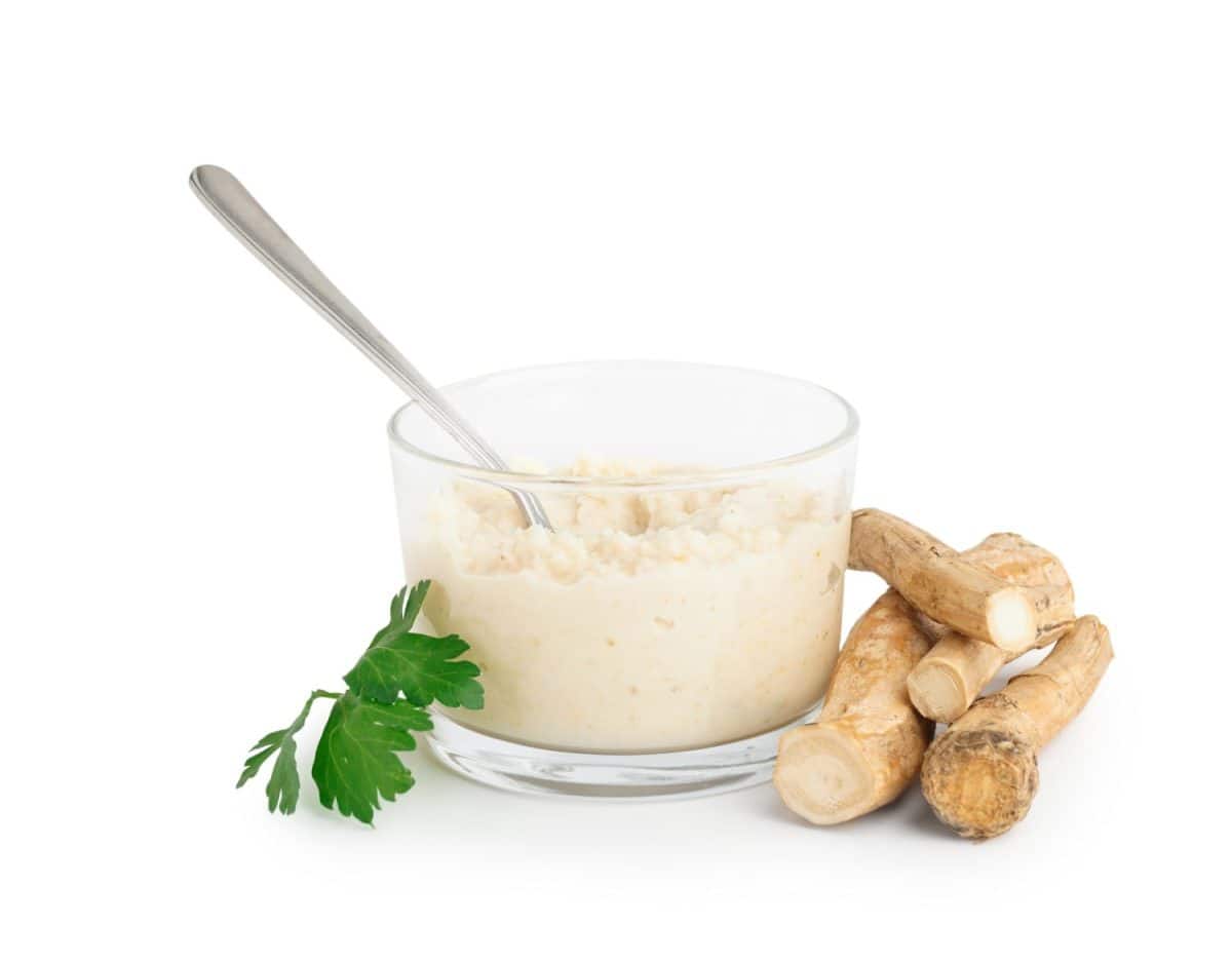 Horseradish sauce in glass bowl with spoon and horseradish root on white background