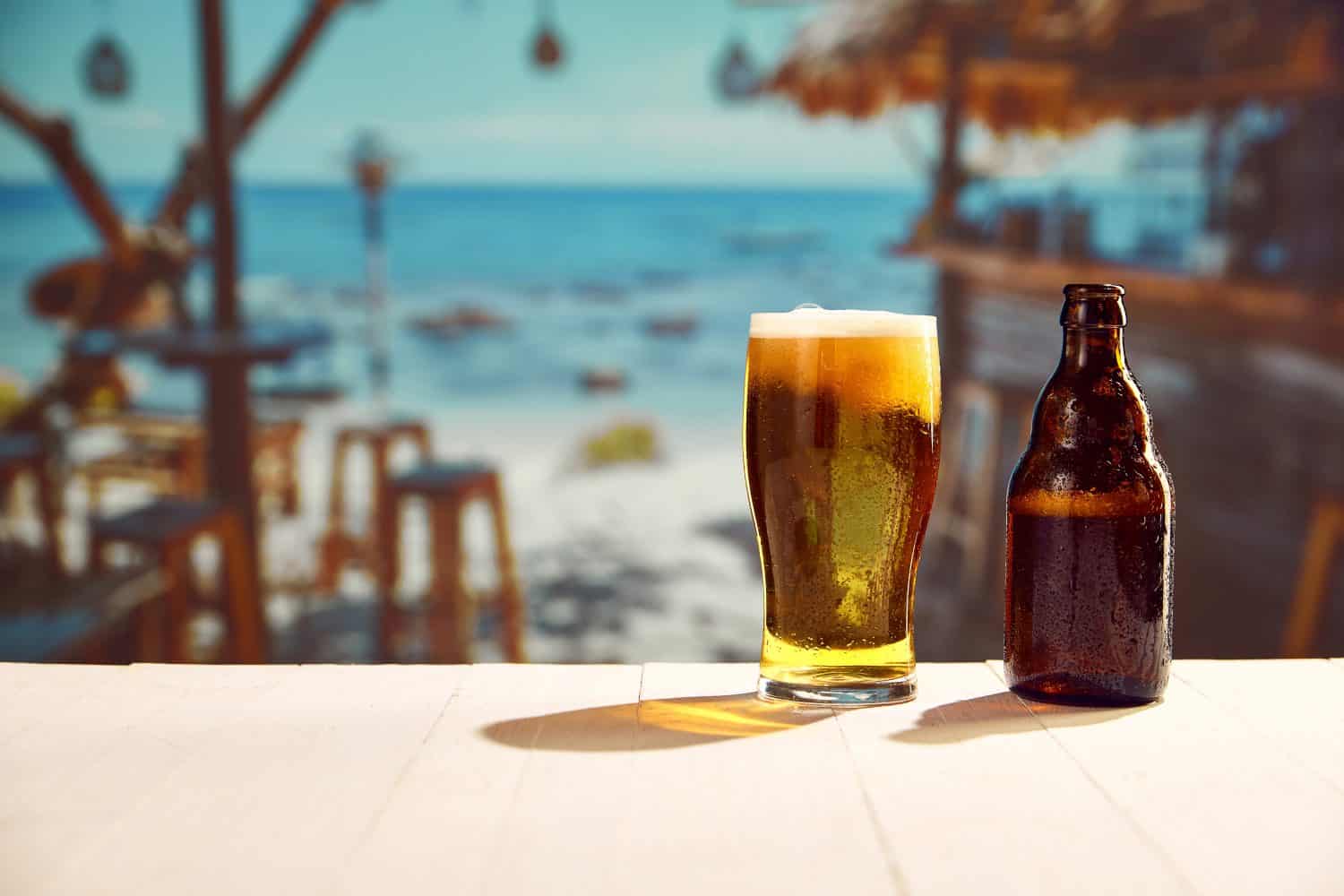 Glass with lager foamy chill beer and beer bottles standing on wooden table at local bar on beach. Sea background. Concept of summer, vacation, relaxation, travelling, leisure time