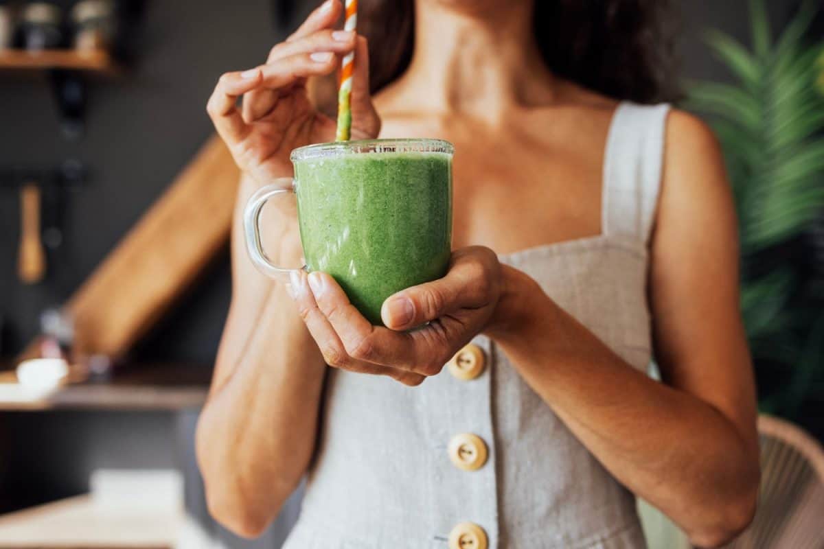 A transparent cup with a green smoothie in the hands of a young woman. A girl in a linen sundress drinks a fresh drink made from herbs and vegetables in the kitchen. Food and drinks are useful.