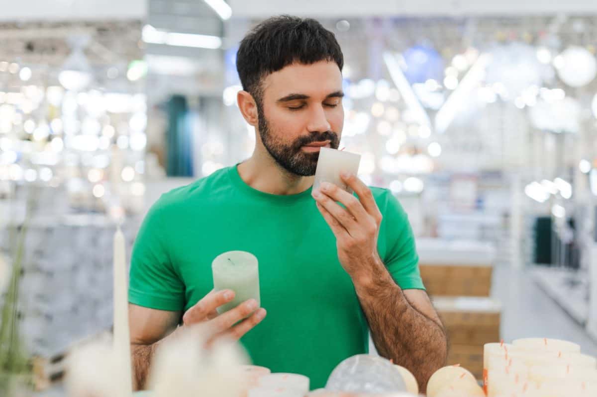 Portrait of young bearded man closing eyes smelling holding decorative candle in a store in the decor department. Shopping concept