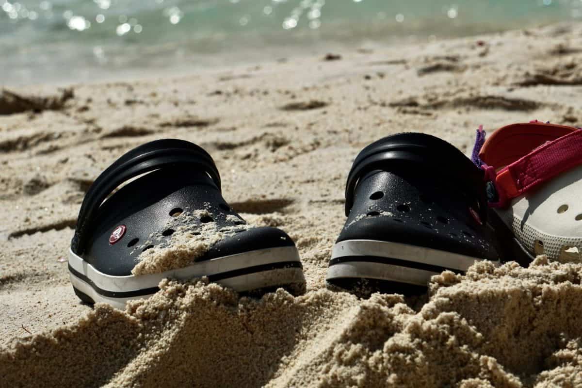 A pair of black and white Crocs clogs partially buried in sand on a beach. The shoes have holes throughout the upper and are covered in sand. The ocean can be seen in the background.