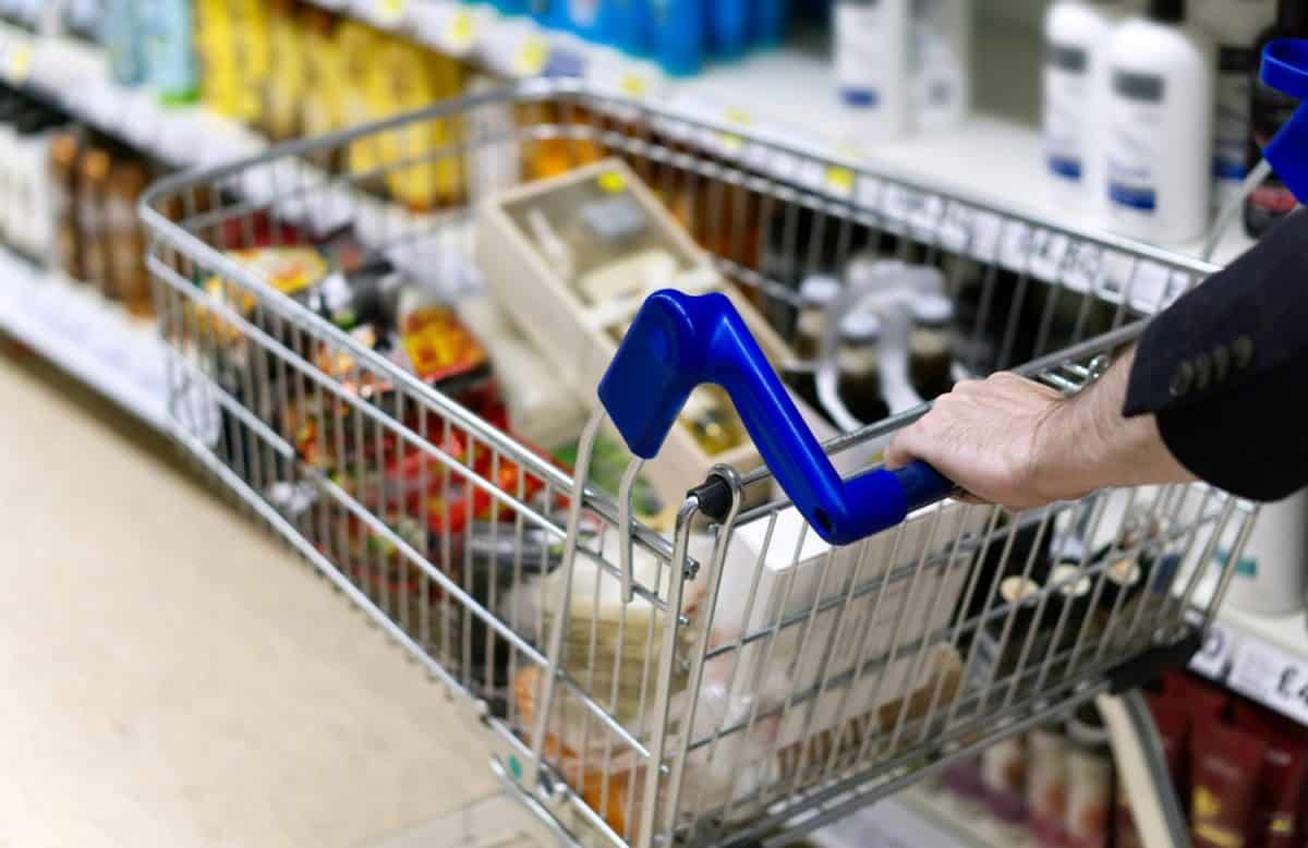 A man's arms pushing a shopping trolley full of groceries along a supermarket aisle.