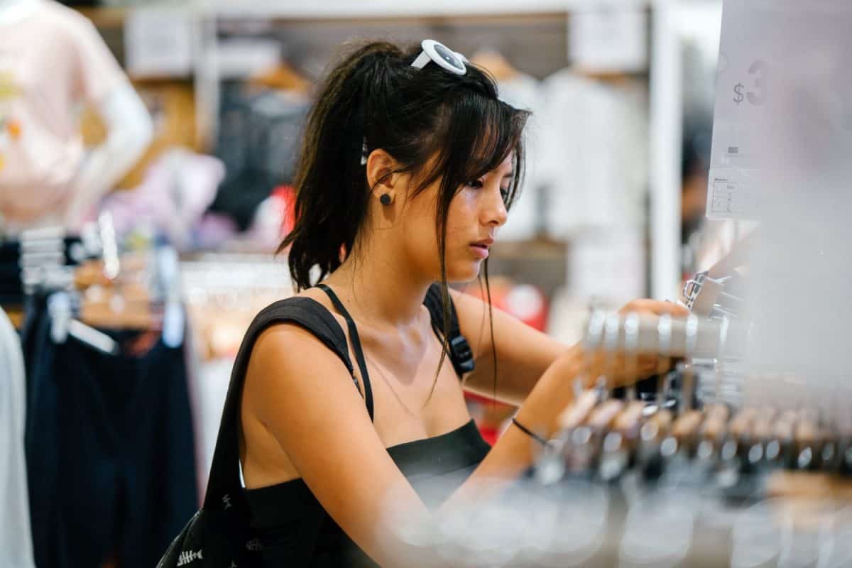 A young and attractive Chinese Asian woman browses the shelves at a store for clothing to buy. The millennial teenager is wearing trendy street clothes and is smiling as she looks through the store.