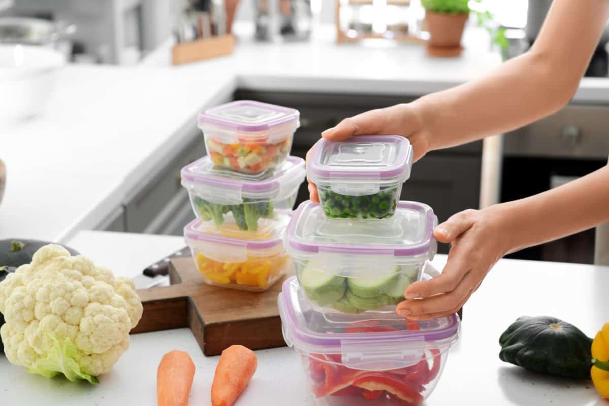 Woman holding stack of plastic containers with fresh vegetables for freezing at table in kitchen