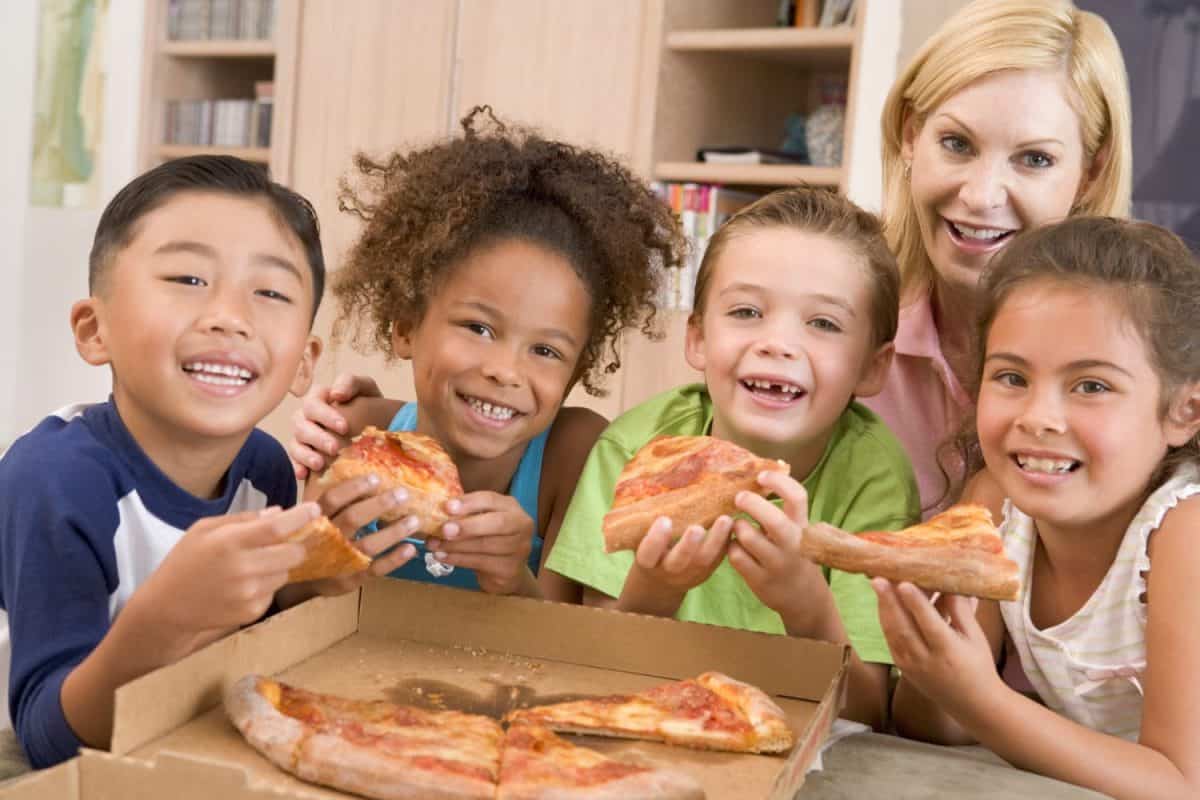 Four young children indoors with woman eating pizza smiling