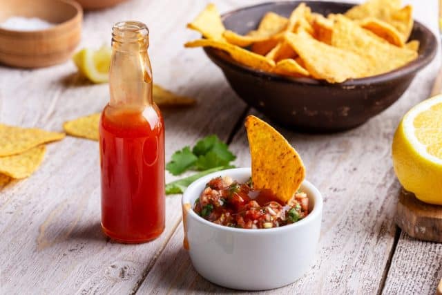 Two bowls full of salsa dip, hot red chili sauce bottle and tortilla chips on rustic wooden table, sause being added to one of the bowls