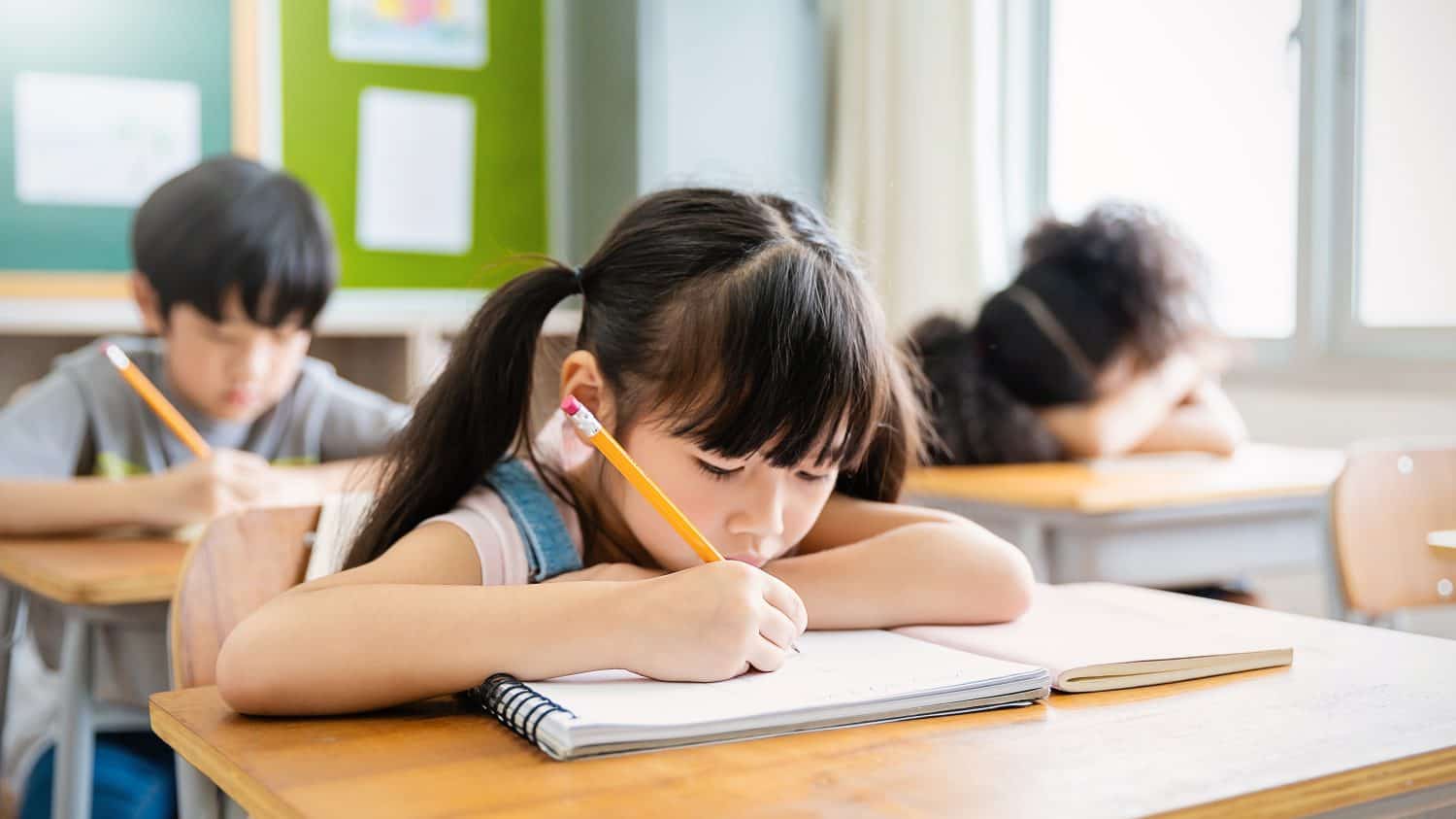 Portrait of little pupil writing at desk in classroom at the elementary school. Student girl study doing test in primary school. Children writing notes in classroom. Education knowledge (blur)