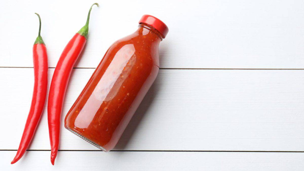 Spicy chili sauce in bottle and peppers on white wooden table, flat lay. Space for text