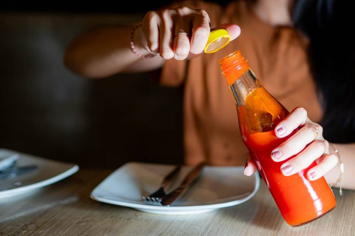 A person opening a bottle of hot sauce at a dining table with plates and cutlery.