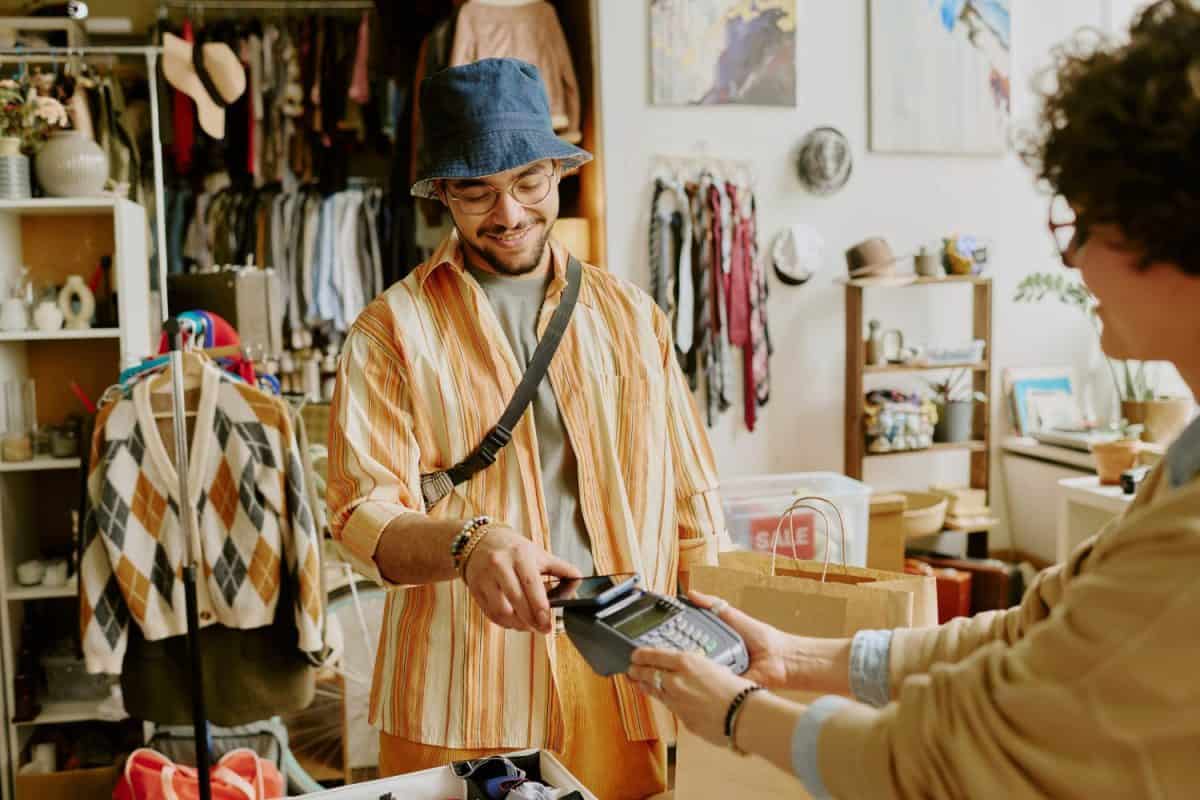 Smiling man wearing hat and glasses making payment at clothing store counter with cashier accepting card. Shelves filled with garments and decorations visible in background