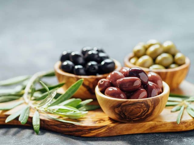 three Woden bowls one each of ripe-black, kalamata, and green olives on a wooden cutting board adorned with rosemary.