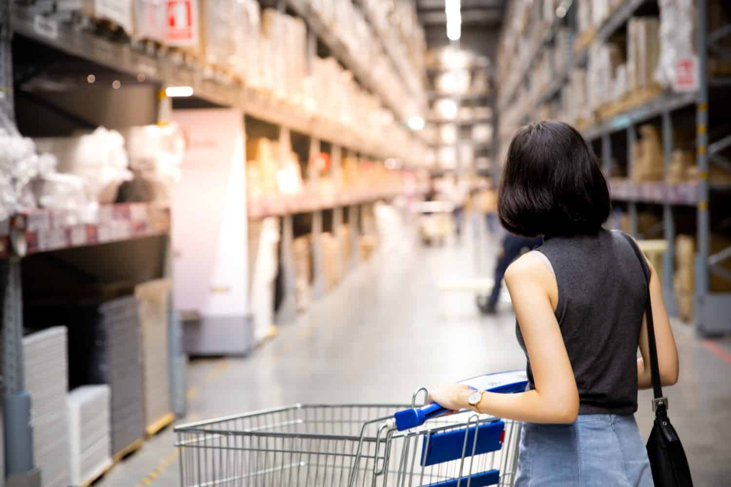 An asian woman doing shopping and walking with her cart in cargo or warehouse. Boxes on rows of shelves in warm light background. copy space