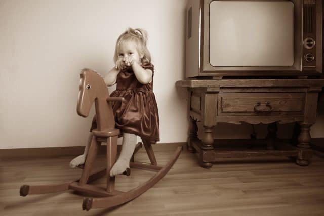 discolored retro style sepia photograph, little blonde girl 2 years old in velvet dress sitting on wooden vintage rocking horse in room by old TV, childhood memories concept