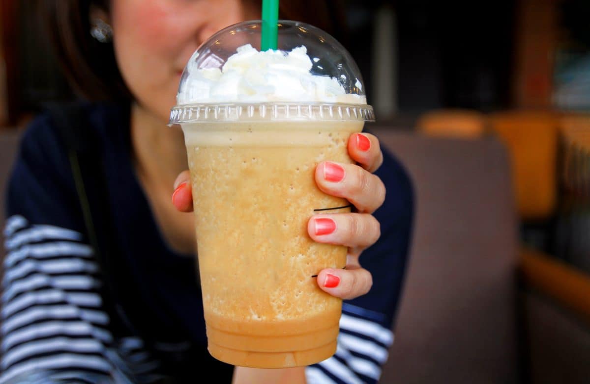 Asian woman in blue casual t-shirt holding a plastic glass of cold espresso Frappuccino with whipped cream on top and her orange nails. The premium coffee. Blended drinking and refreshment concept.