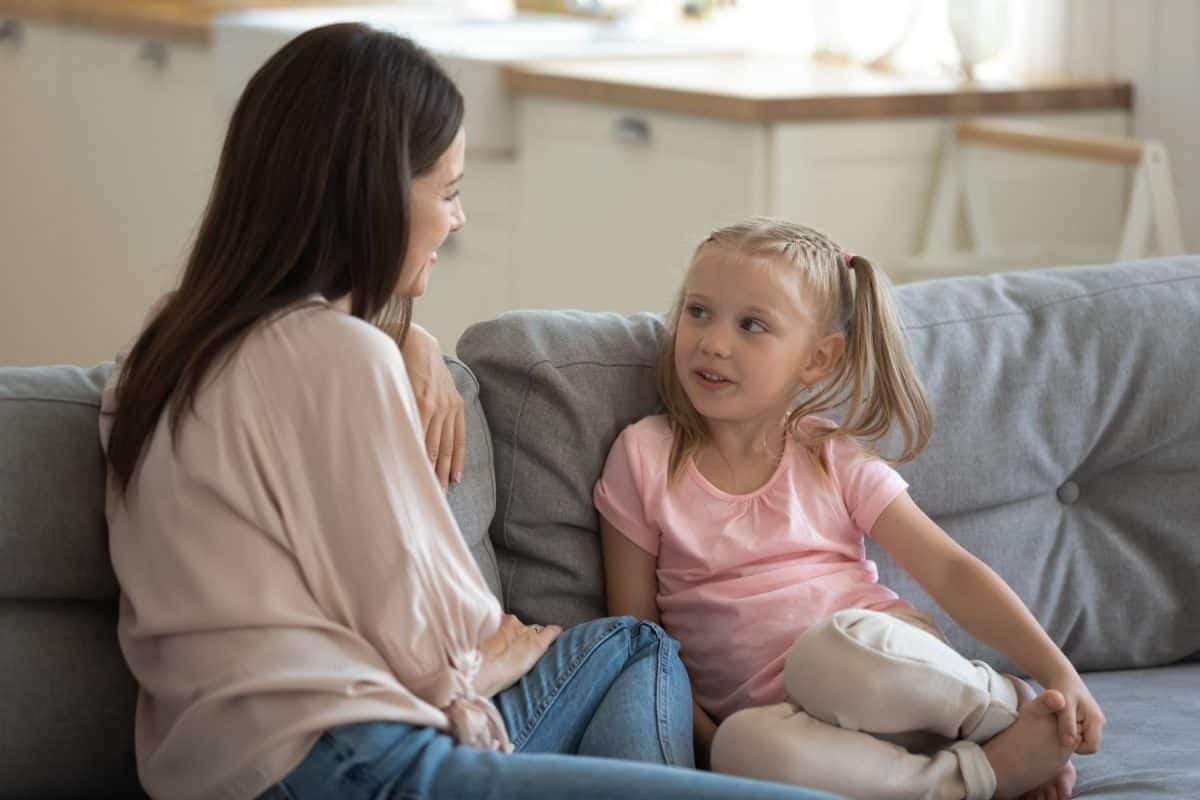 Happy mother and little daughter talking, sharing news, sitting on comfortable couch in living room, young mum and adorable preschool girl chatting, family spending weekend together at home