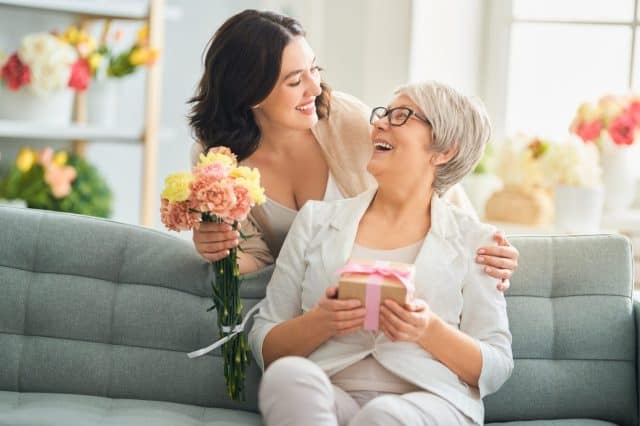 Happy mother's day! Beautiful young woman and her mother with flowers and gift box at home.