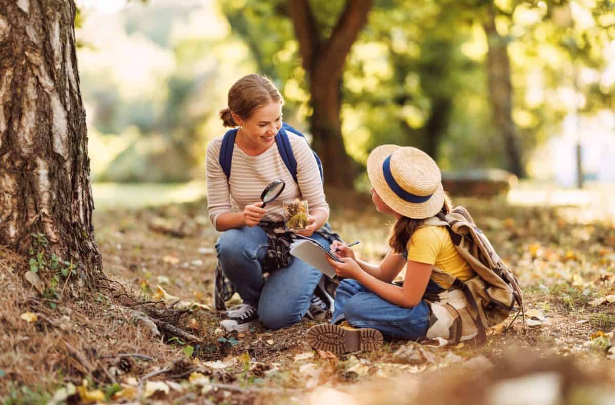 Child girl and female teacher with backpacks looking examining plants and insects in a jar through magnifying glass while exploring forest nature and environment on sunny day during outdoor ecology