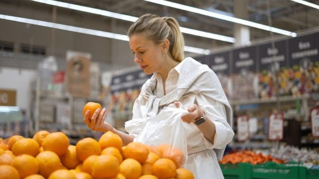 Woman buy healthy food fruit market. Customer take ripe orange eco store. 30s lady keep raw diet. Girl go shop bio mall. Person put many item bag. Vegan meal row. Health care. Pick up vitamin c juice.