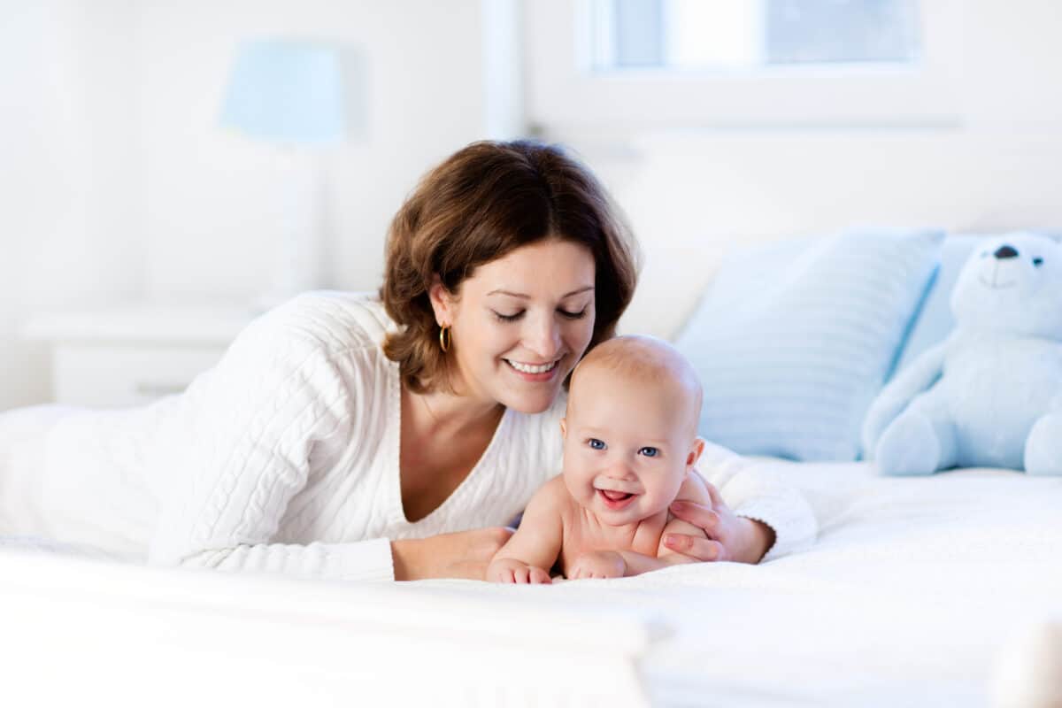 Mother and child on a white bed. Mom and baby boy in diaper playing in sunny bedroom.